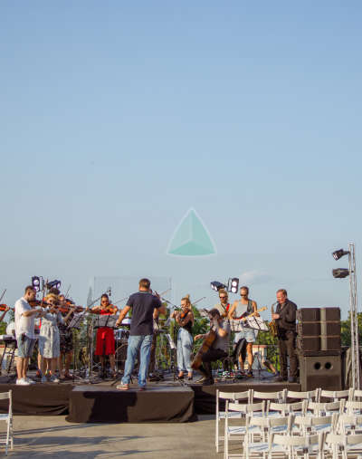 Un grupo de músicos actúa en un escenario al aire libre con un director de orquesta, mientras unas sillas blancas vacías se disponen frente al escenario bajo un cielo despejado.