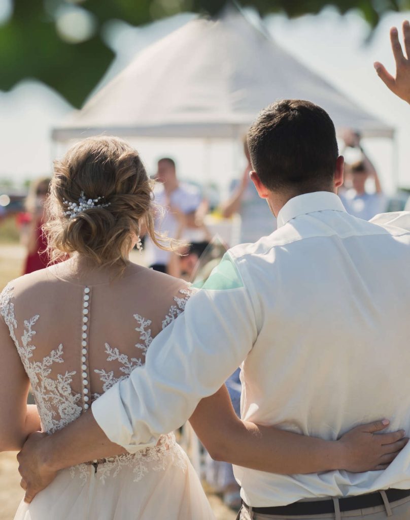 Una pareja de novios se rodea con los brazos, de espaldas a la cámara, mientras el novio saluda con la mano a un grupo de personas en un evento al aire libre.