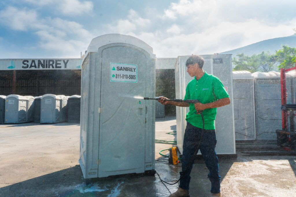 Un trabajador con una camiseta verde utiliza una hidrolimpiadora para limpiar un baño portátil al aire libre en un día soleado.