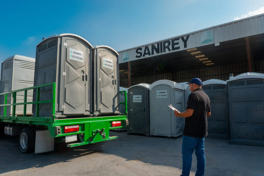 Un hombre revisa unos baños portátiles junto a un camión verde en unas instalaciones con el letrero «Sanirey» en un día soleado.