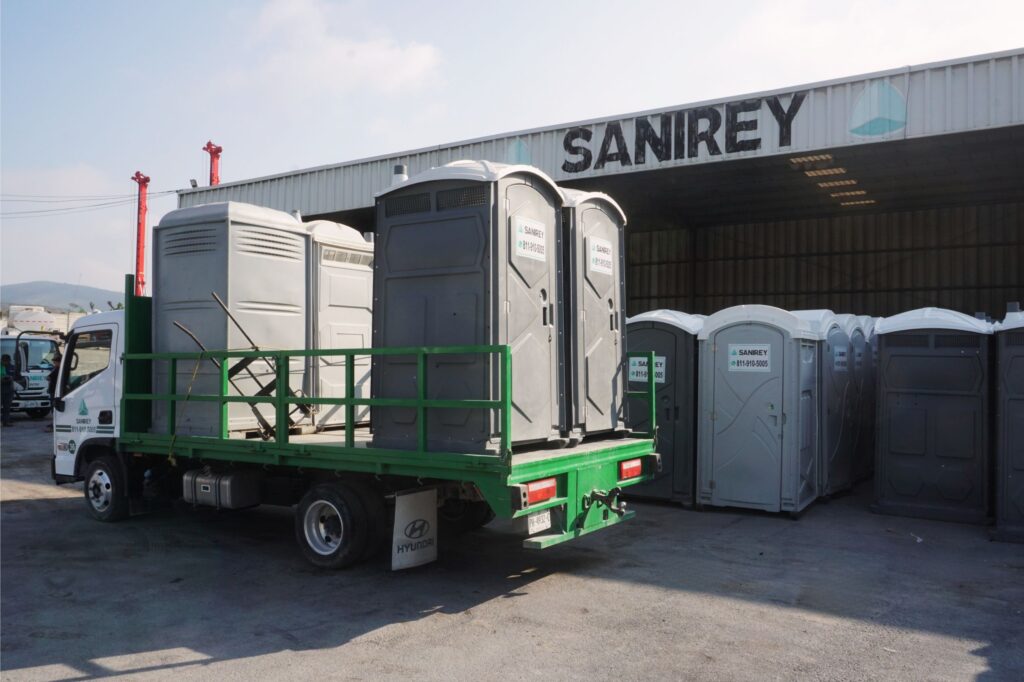 Un camión de plataforma cargado con baños portátiles está aparcado frente a un almacén con el letrero «SANIREY», y cerca hay más baños portátiles apilados.