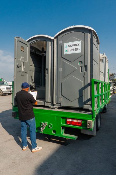 Un trabajador está junto a un camión verde cargado de baños portátiles; sostiene un portapapeles y revisa las unidades bajo un cielo azul y despejado.