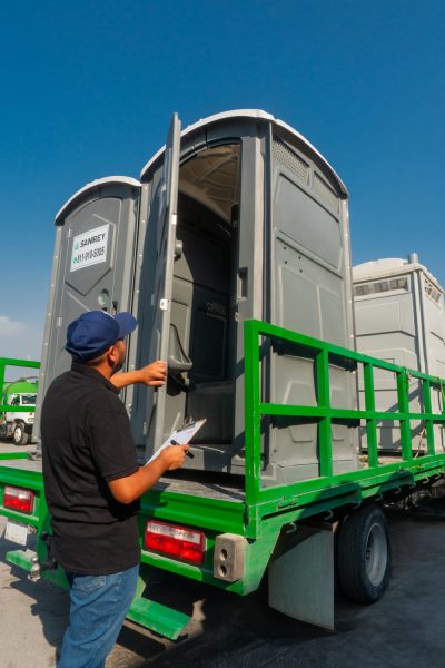 Un hombre con una camiseta negra inspecciona un baño portátil en un camión de plataforma verde, con un portapapeles en la mano, bajo un cielo azul claro.